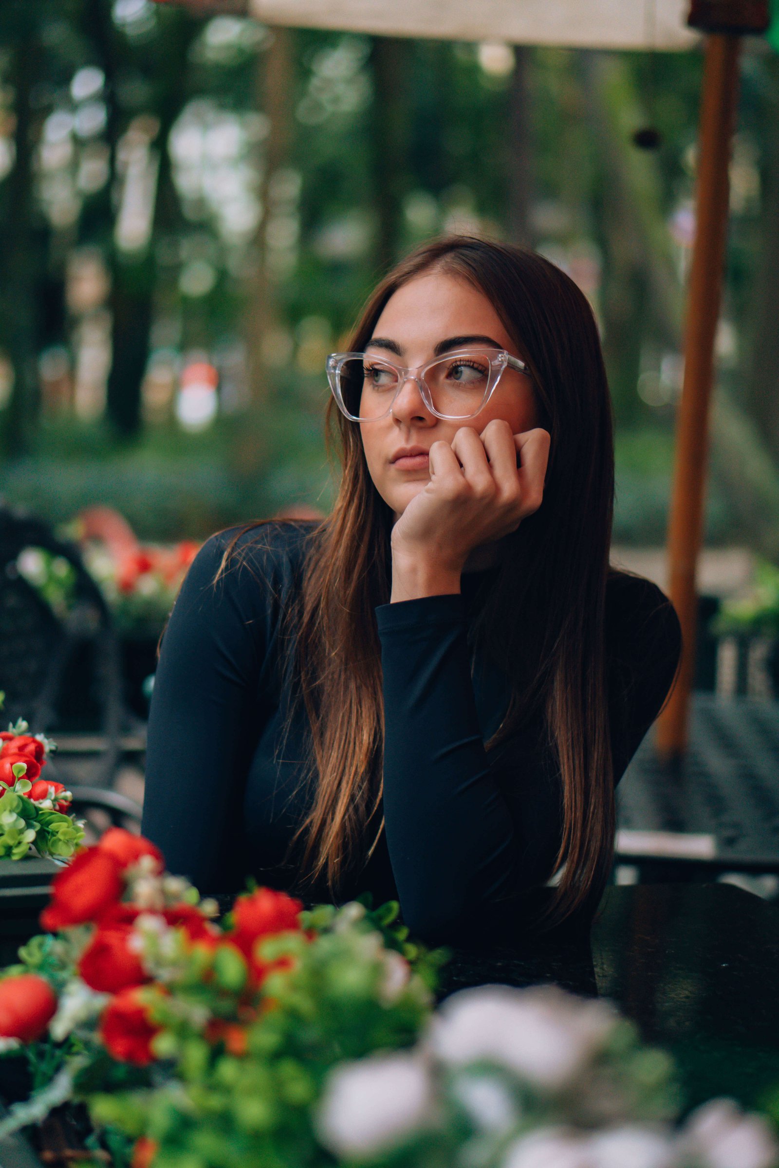 Mulher com óculos de grau e cabelos longos posando ao ar livre ao lado de vasos com flores vermelhas.