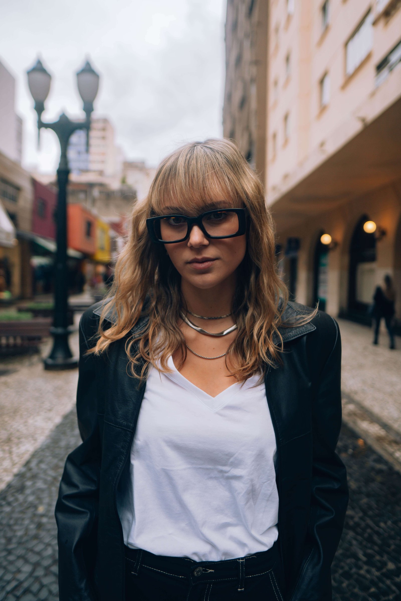 Mulher com franja e óculos de grau em rua de Curitiba, usando blazer e camisa branca.