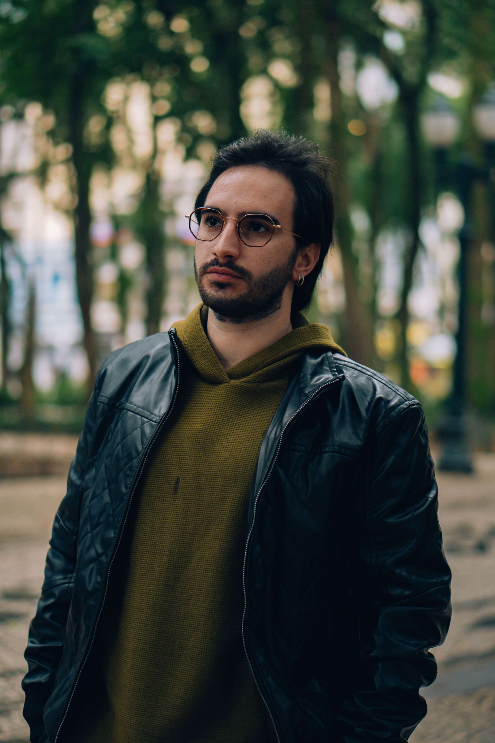 Homem com óculos de grau, barba e jaqueta de couro caminhando em parque arborizado.