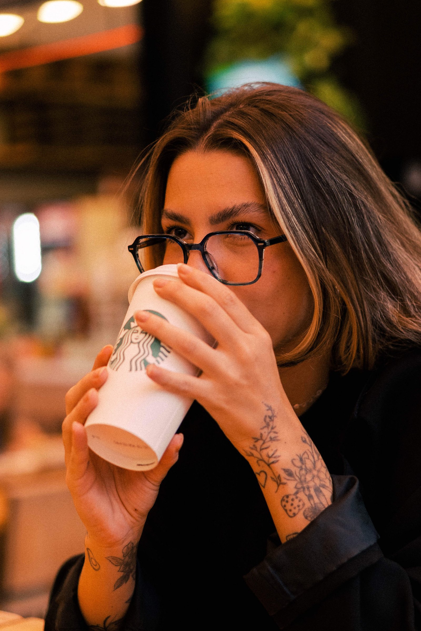 Mulher usando óculos de grau bebendo bebida gelada com canudo em cafeteria iluminada.