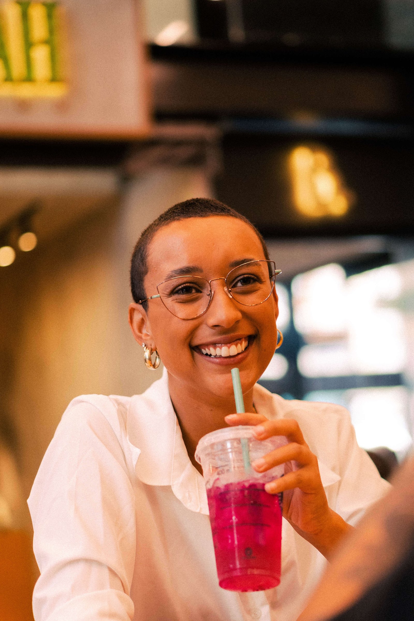 Mulher sorridente de cabelos curtos usando óculos de grau e segurando copo com bebida rosa em cafeteria.