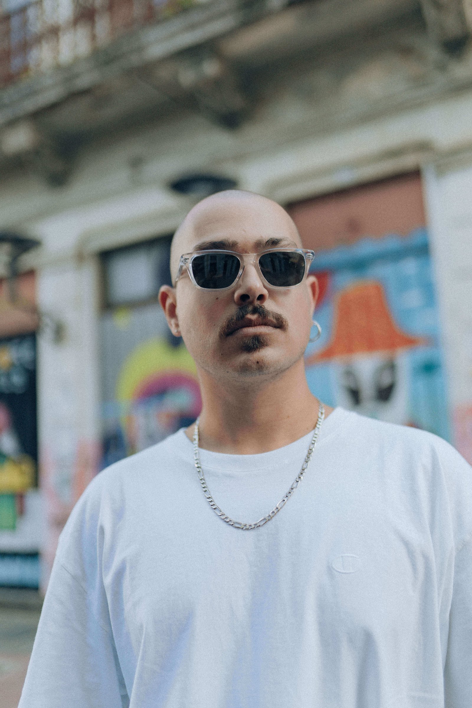 Homem usando óculos de sol escuros e camiseta branca posando em rua com grafites ao fundo.