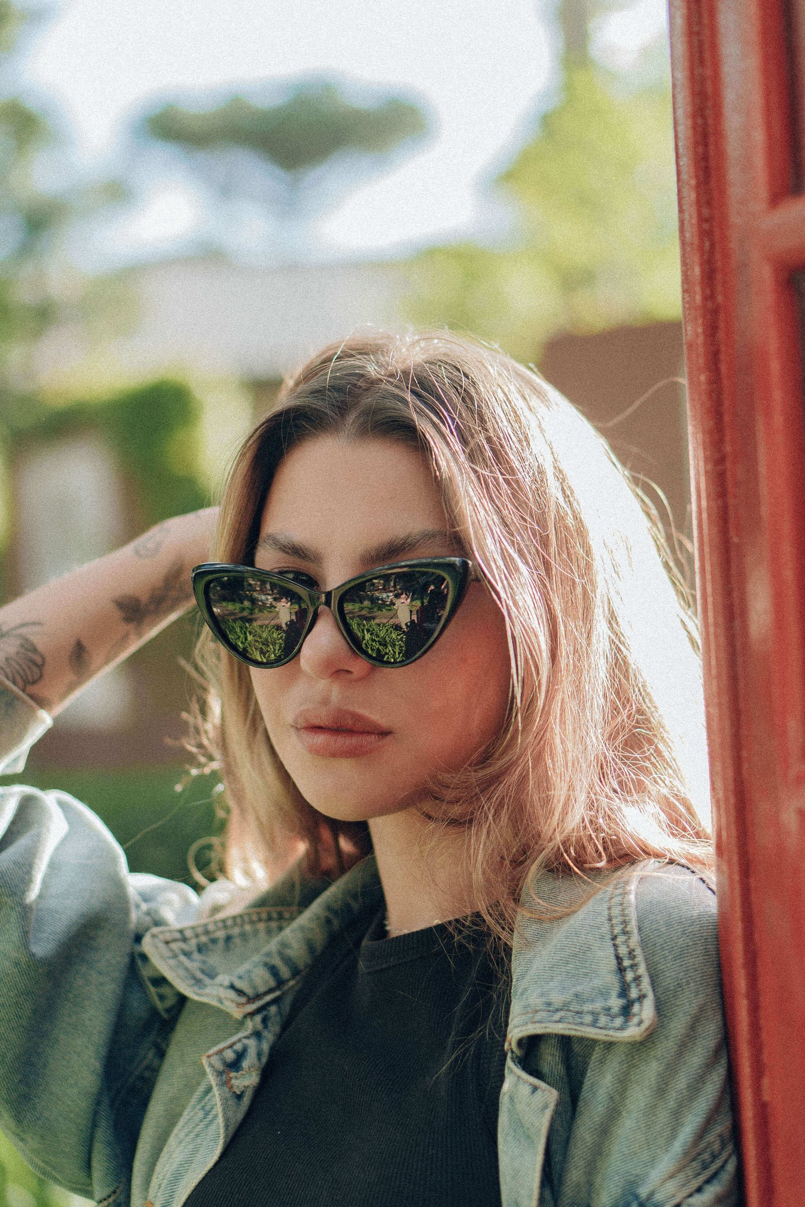 Mulher de cabelo claro usando óculos de sol com lentes espelhadas verdes e jaqueta jeans em área externa arborizada.
