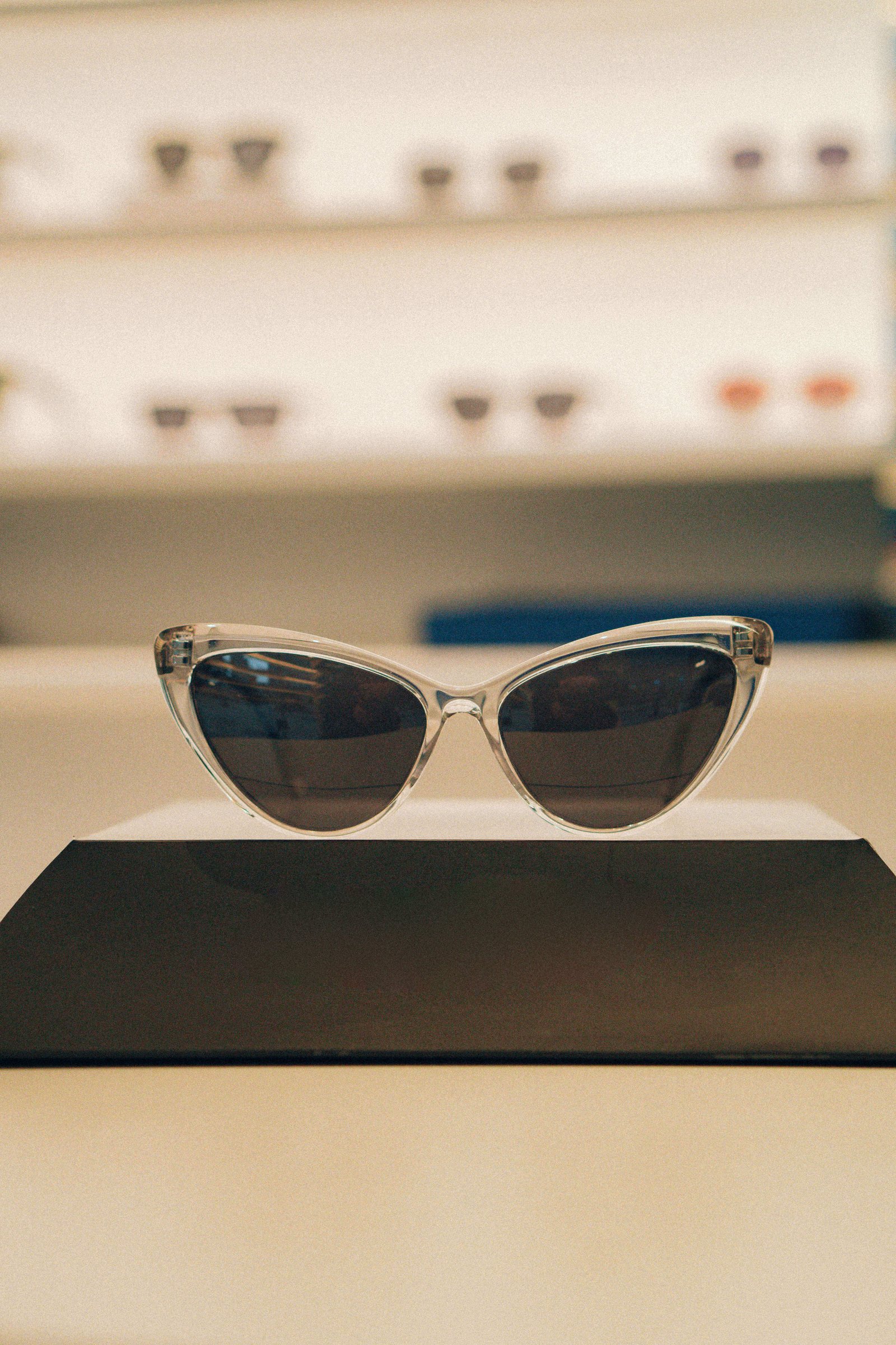 Óculos de sol pretos em formato gatinho apoiados em base preta com fundo de vitrine desfocado.