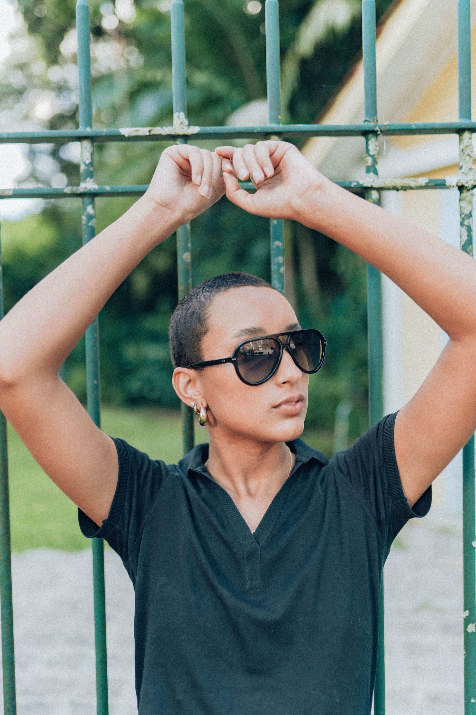 Pessoa com óculos de sol grandes e camiseta preta apoiando os braços na grade de um parque.