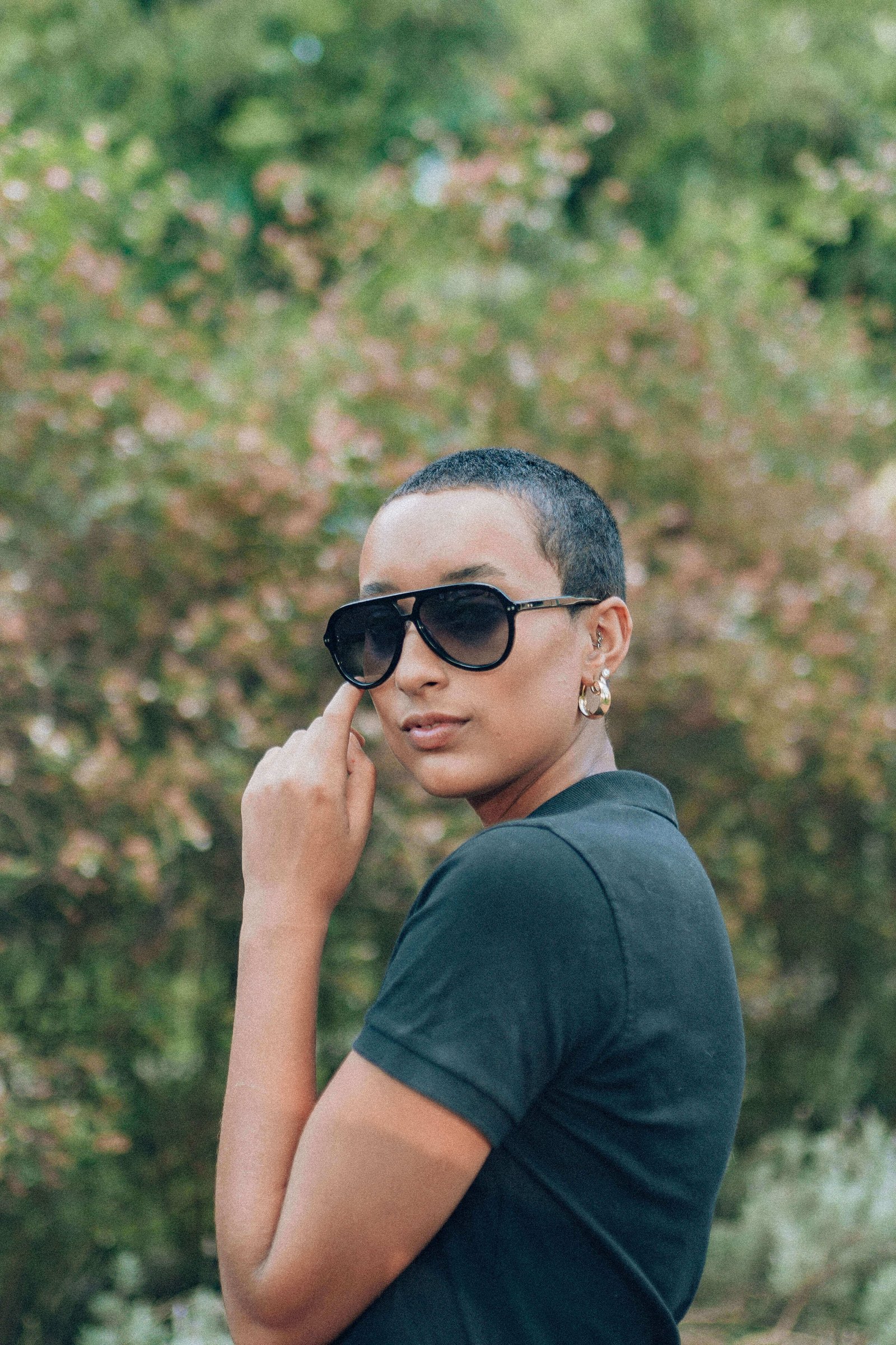 Pessoa de perfil usando óculos de sol quadrados pretos em área externa com vegetação ao fundo.