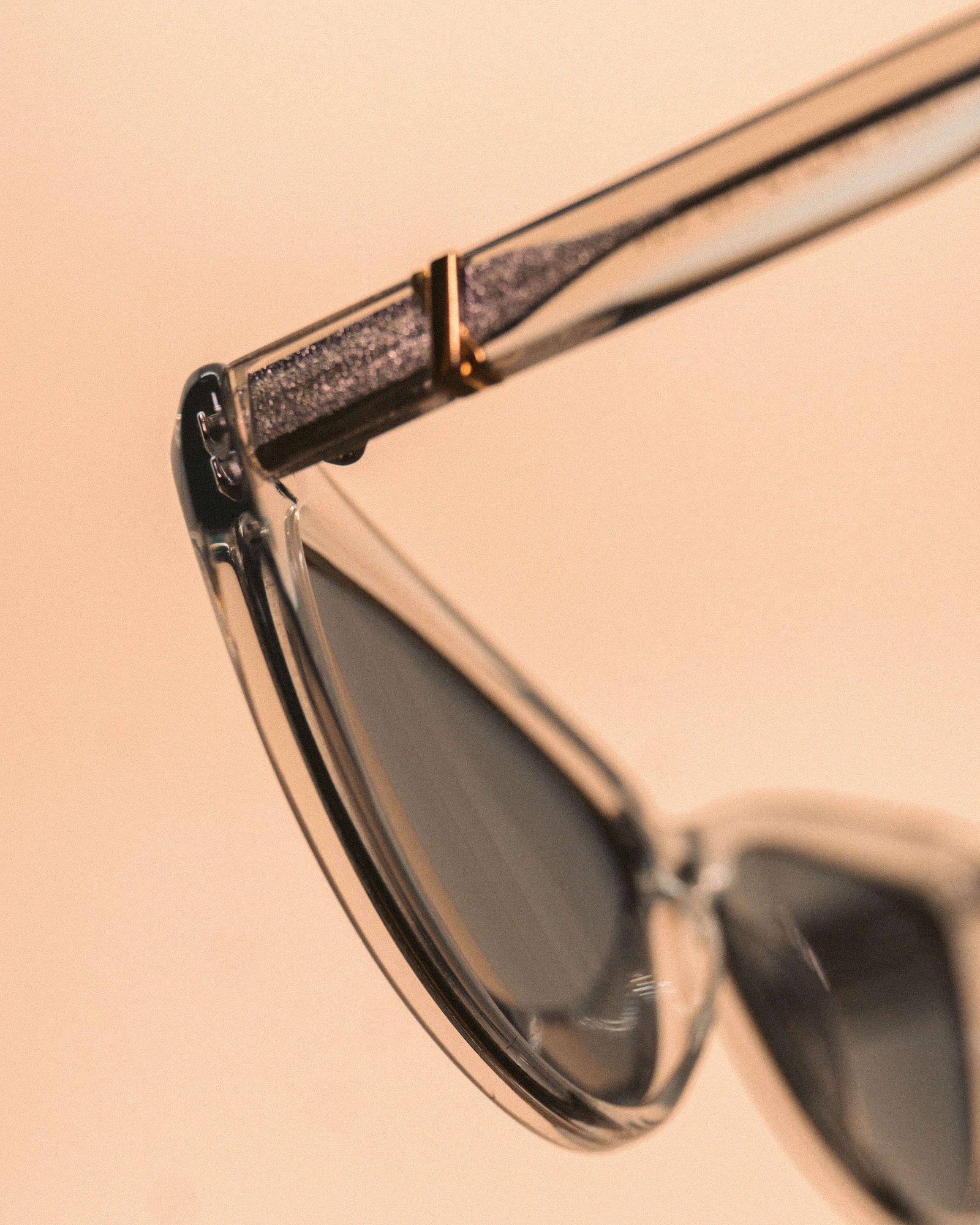 Close de um óculos de sol estilo gatinho em metal prateado com lentes escuras sobre fundo bege, destacando o detalhe da haste.